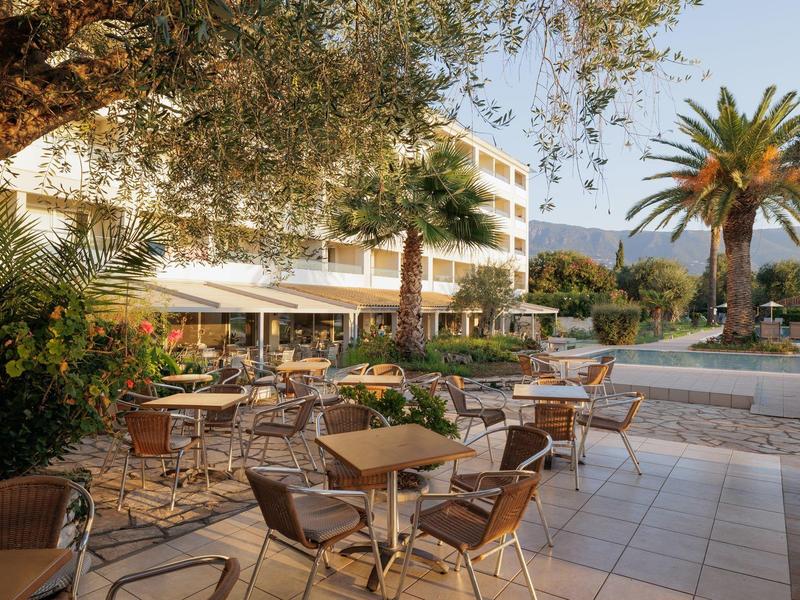 Terrassencafé mit Tischen und Stühlen umgeben von Palmen und Bäumen, im Hintergrund mehrstöckiges Gebäude.