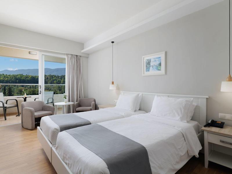 Helles Hotelzimmer mit zwei Betten, grauen Decken, Sesseln und Balkon mit Bergblick.