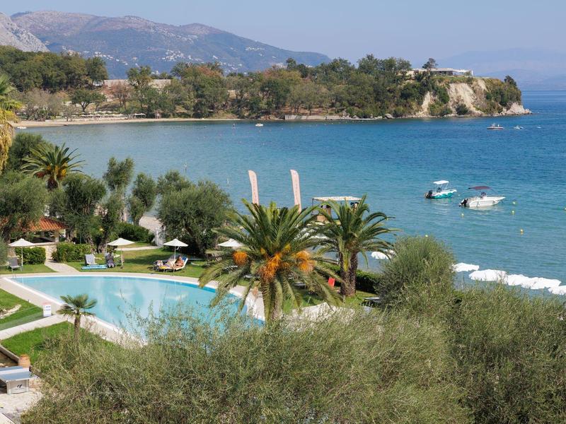 Blick auf einen blauen Pool, Palmen und Boote im türkisfarbenen Meer mit Bergen im Hintergrund.