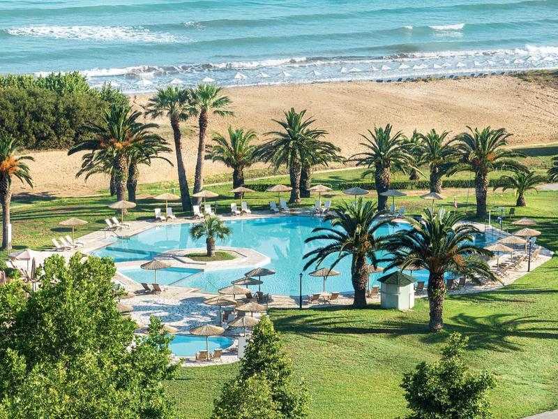 Blick auf einen blauen Pool mit Palmen, Liegestühlen und einem Sandstrand im Hintergrund.