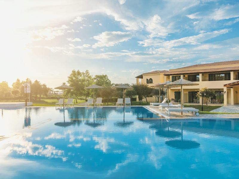 Großer, klarer Pool mit Liegestühlen, umgeben von Gebäuden und Bäumen bei Sonnenschein.