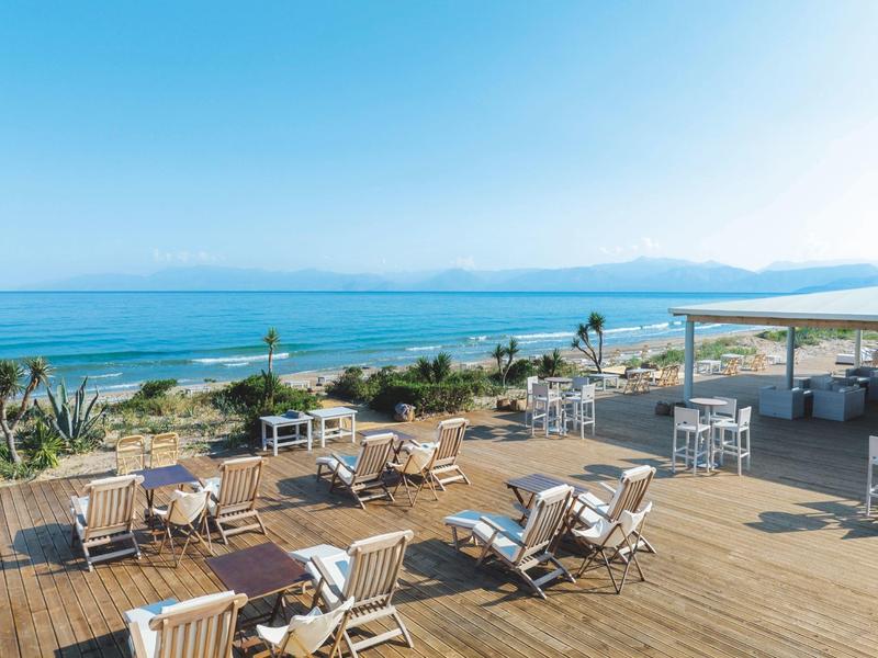 Große Holzterrasse mit weißen Liegestühlen und Tischen, Blick auf Meer und klaren Himmel.