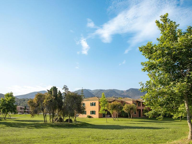 Grüner Park mit Bäumen, Wiese, gelbem Gebäude und Bergen im Hintergrund unter blauem Himmel.
