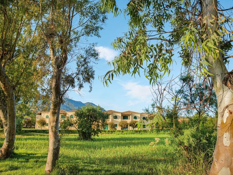 Grüne Wiese mit Bäumen im Vordergrund und gelbliche Gebäude mit Bergen im Hintergrund unter blauem Himmel.