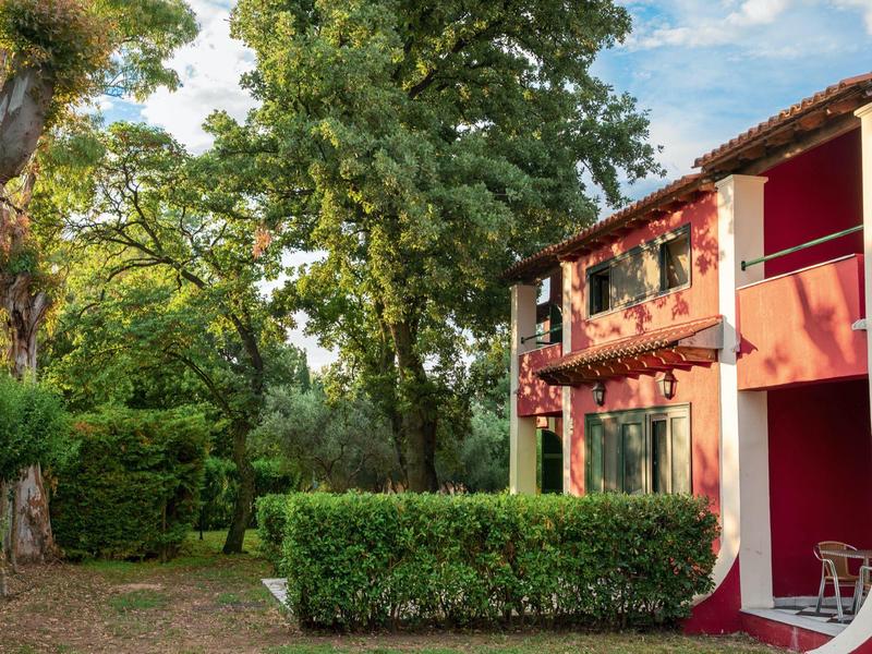 Haus mit roter Fassade, grünen Hecken und großen Bäumen unter blauem Himmel an sonnigem Tag.