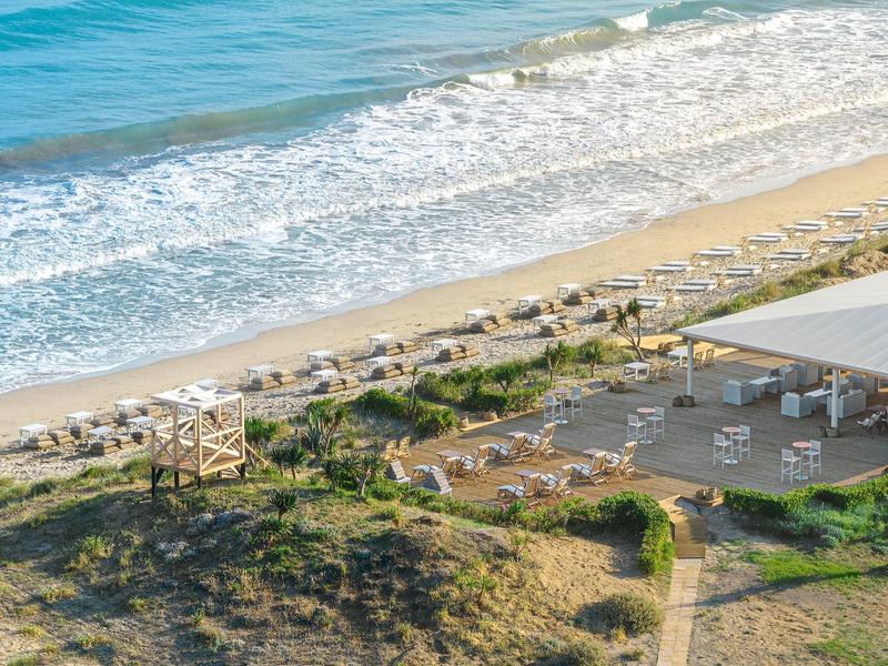 Strand mit Sand, Wellen, Liegen, Sonnenschirmen und einem großen überdachten Bereich am Ufer.