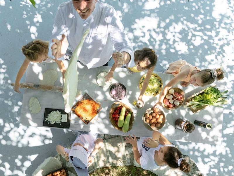Gruppe von Menschen sitzt an Tisch im Freien, isst, trinkt und genießt sonnigen Tag mit schattigem Laubdach.