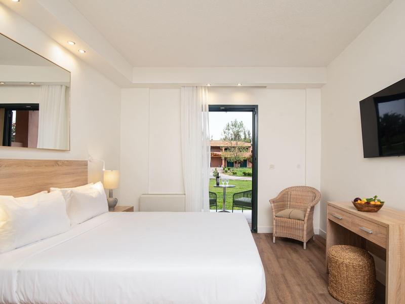 Helles Schlafzimmer mit großem Bett, Sessel, TV an der Wand und Blick auf einen Garten durch die Tür.