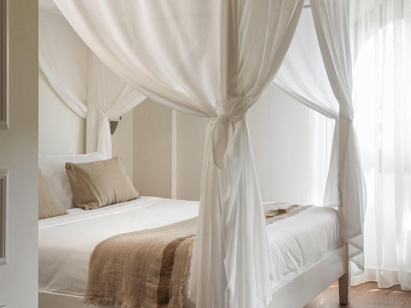 Helles Schlafzimmer mit weißem Bett, beigen Kissen und transparenten Vorhängen am Fenster.