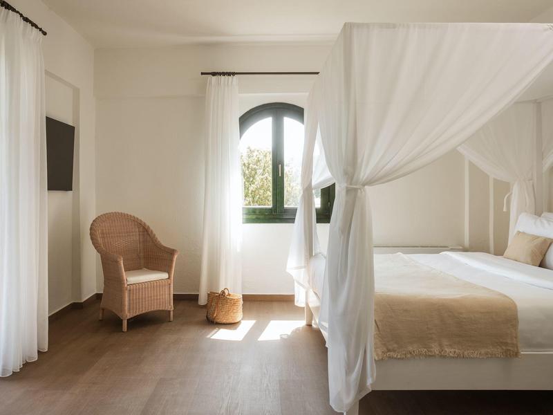 Helles Schlafzimmer mit Himmelbett, Rattanstuhl, Bogenfenster und weißen Vorhängen.