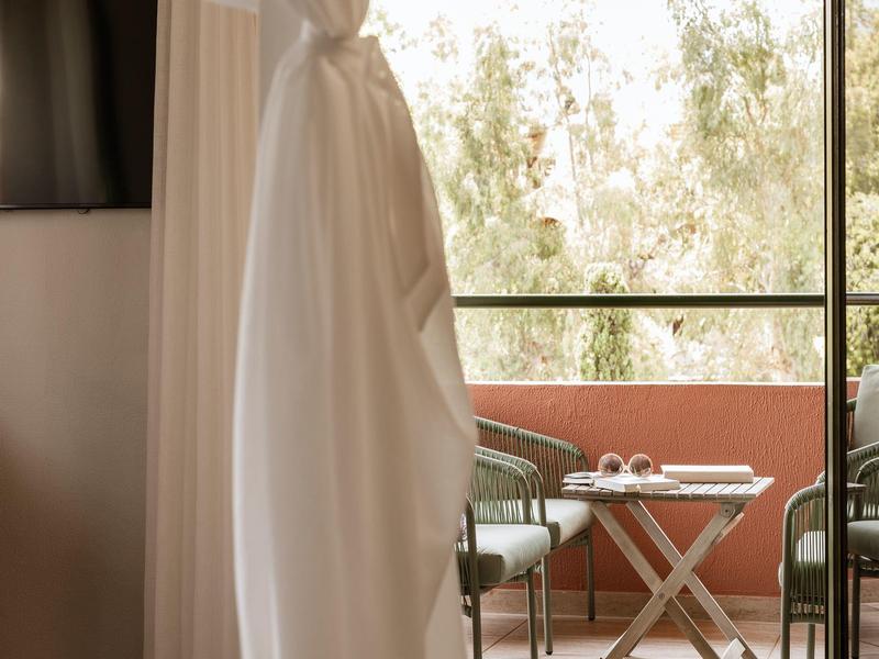 Balkon mit grünen Stühlen, kleinem Tisch und weißen Kissen, vor hellen Vorhängen und Bäumen.