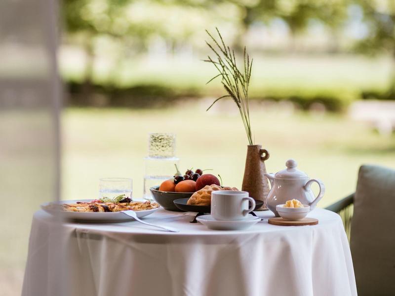Tisch mit weißer Decke, Teekanne, Tassen, Obstschale und Vase mit Gräsern auf Terrasse.