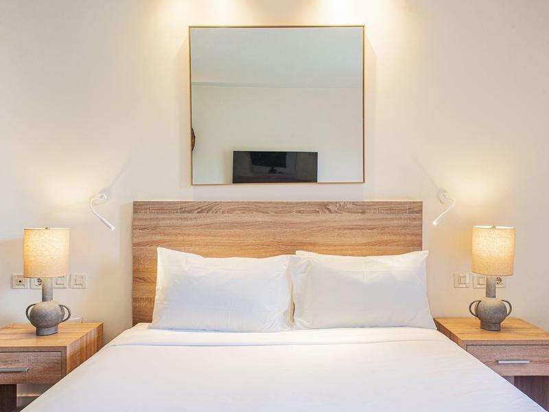 Hell eingerichtetes Schlafzimmer mit Holzbettrahmen, weißen Kissen, zwei Nachttischlampen und großem Spiegel.