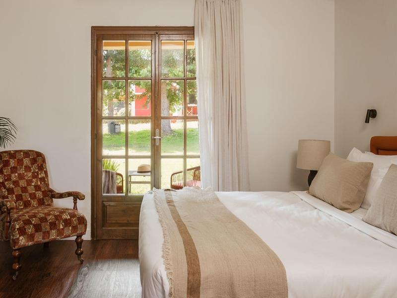 Helles Schlafzimmer mit Holzfenster, weißer Bettwäsche, beigem Bettläufer und braun gemusterter Sitzbank.