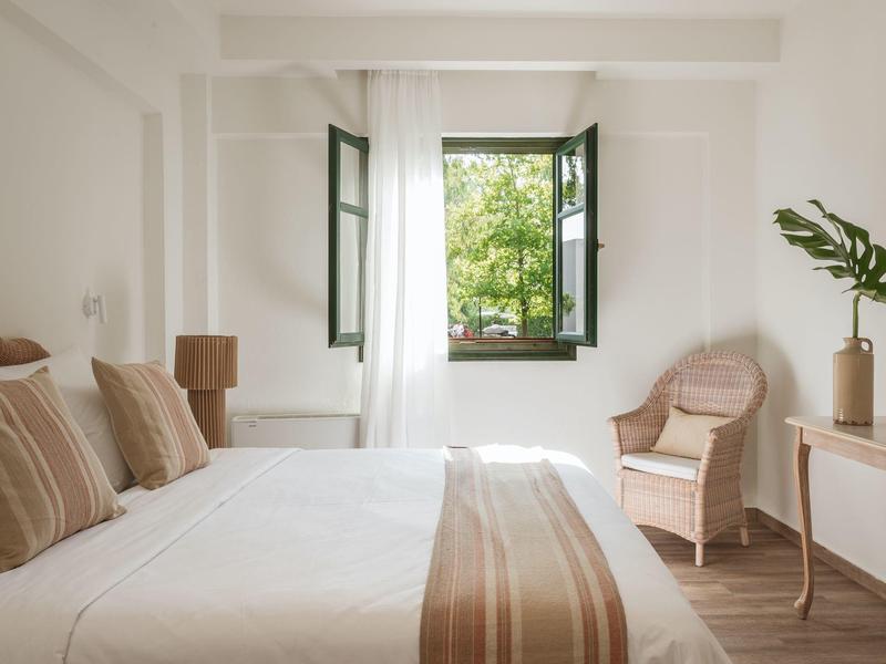 Helles Schlafzimmer mit offenem Fenster, weißer Bettwäsche, gestreiften Kissen und Holzstuhl.