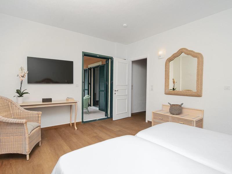 Schlichtes Schlafzimmer mit weißen Wänden, Holzboden, Doppelbett, Korbstuhl, Spiegel und TV an der Wand.