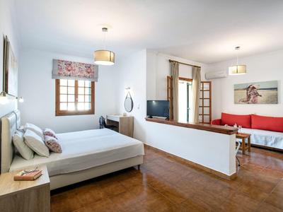 Helles Zimmer mit Bett, Sofa, Fernseher, Holzboden und großen Fenstern mit weißen Wänden.