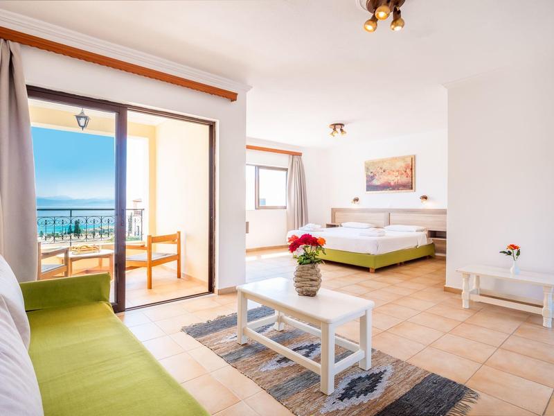 Bright hotel room with sea view balcony, bed, sofa, coffee table, and tiled floor.