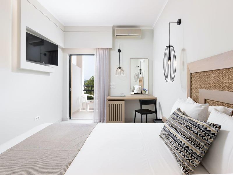 Hell eingerichtetes Schlafzimmer mit Doppelbett, Schreibtisch, Stuhl und Balkon mit Blick nach draußen.