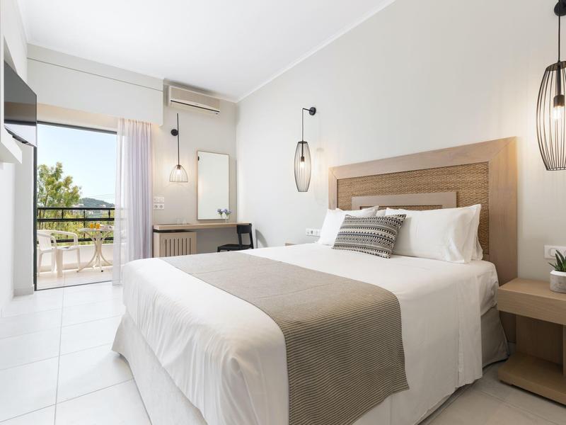 Helles Schlafzimmer mit Doppelbett, grauem Bettläufer, Nachttisch und großem Fenster mit Blick ins Grüne.