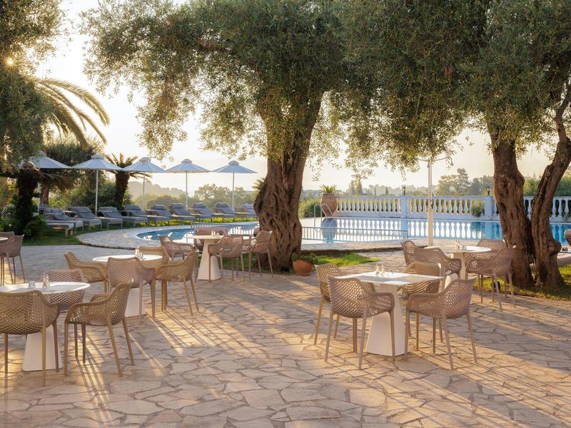 Terrasse mit Stühlen und Tischen unter großen Bäumen neben Pool mit Sonnenschirmen und Garten.