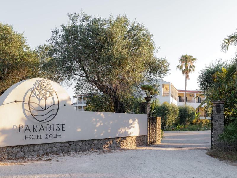 Eingang zu Paradise Home Goa mit weißer Mauer, Logo, Pflasterweg und umliegenden Bäumen.