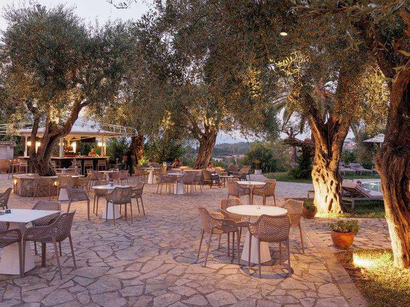 Leerer Außenbereich eines Cafés mit runden Tischen und Stühlen auf Steinboden unter Olivenbäumen.