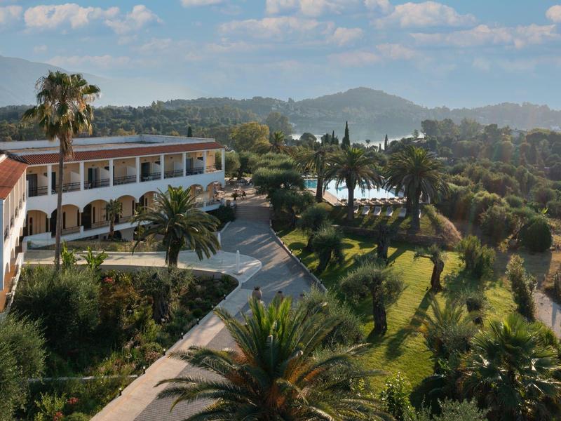 Hotelanlage mit weißen Gebäuden, Palmen und grünen Rasenflächen vor bewaldeten Hügeln unter blauem Himmel.