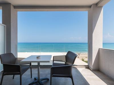 Balkon mit zwei schwarzen Stühlen und weißem Tisch, Blick auf ruhiges blaues Meer und Himmel.