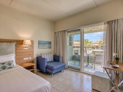 Modernes Hotelzimmer mit Bett, Sofa, Schreibtisch und Terrasse mit Blick auf Palmen.