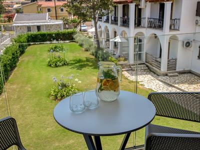 Blick von einem Balkon auf einen Garten mit Rasen, Blumenbeeten und einem runden Tisch mit Karaffe und Gläsern.