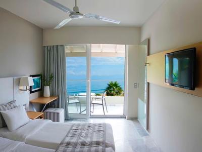 Helles, modernes Zimmer mit zwei Betten, Fernseher, Deckenventilator und Meerblick durch große Balkontür.