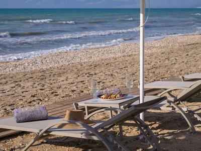 Mehrere Liegestühle mit Handtüchern und Snacks am Sandstrand neben dem Meer unter blauem Himmel.