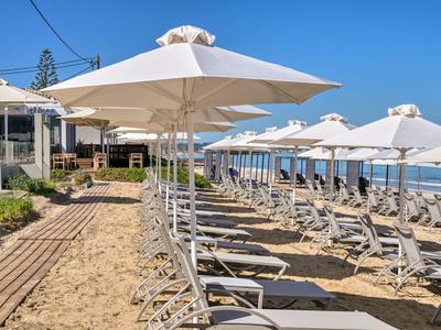 Reihe von weißen Liegestühlen und Sonnenschirmen am Sandstrand neben einem Hotel bei klarem Himmel.