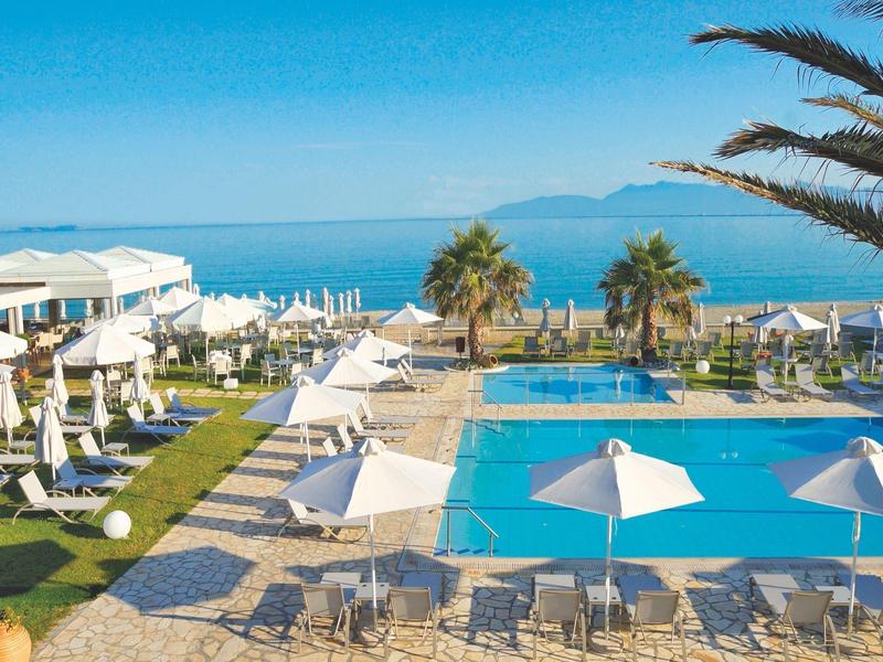 Hotelpool mit Sonnenliegen, Schirmen und Meerblick unter klarem Himmel.