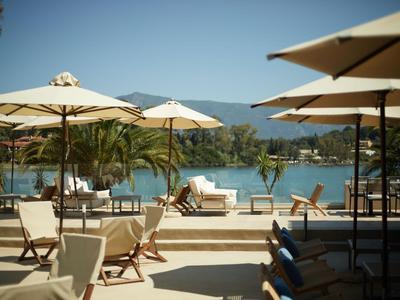 Poolbereich mit weißen Sonnenschirmen und Stühlen, Palmen und Blick auf See und Berge.