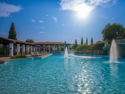 Großer Außenpool mit Wasserfontänen und Sonnenliegen, umgeben von Gebäuden und Bäumen.