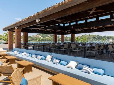 Offener Dachbereich mit blauen Sofas und Holztischen an einem Hotelpool mit Blick auf Hügel.