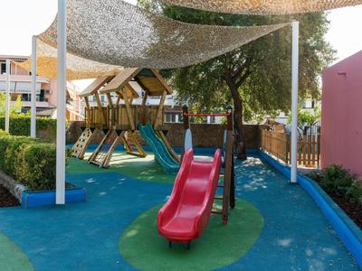 Spielplatz mit bunter Rutsche und Schattensegeln neben einem Hotelgebäude.