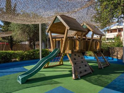 Spielplatz mit Holzhaus, Rutsche und Kletterwand auf grünem Boden unter Sonnennetz