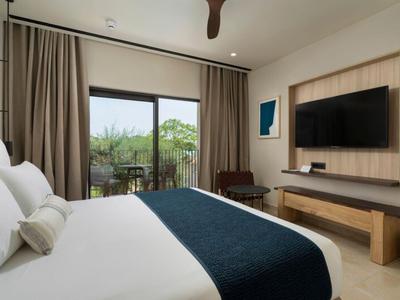 Hell eingerichtetes Hotelzimmer mit großem Bett, Fernseher an der Wand und Blick auf Balkon mit grüner Landschaft.