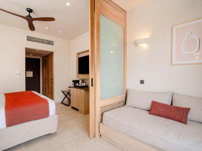 Helles Hotelzimmer mit Doppelbett, grauem Sofa, Holzdetails und Deckenventilator.