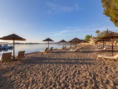 Ein ruhiger Sandstrand mit Sonnenschirmen und Liegestühlen neben klarem, ruhigem Wasser unter blauem Himmel.