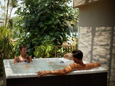 Zwei Personen entspannen in einem Whirlpool auf einer Terrasse mit Pflanzen im Hintergrund.