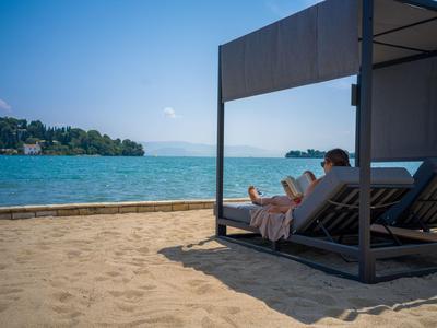 Zwei Personen entspannen auf Liegestühlen unter einem Sonnenschutz am Sandstrand mit Blick auf das Meer.