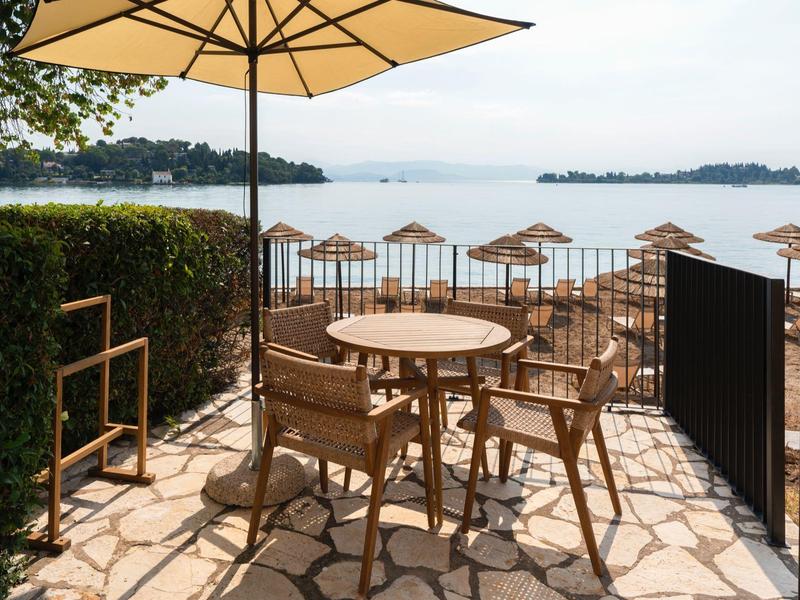 Terrassenbereich mit Holztisch, Stühlen und Sonnenschirm am Seeufer mit Sandstrand und Schirmen.