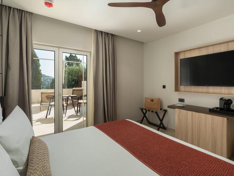 Hell eingerichtetes Schlafzimmer mit großem Bett, Fernseher, Deckenventilator und Zugang zur Terrasse.