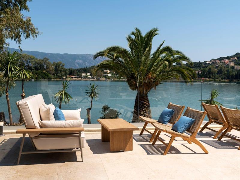 Terrasse mit Holzliegen, blauen Kissen und Palmen am Meer bei klarem Himmel.