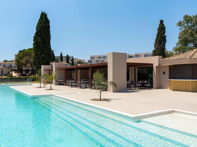 Ein großer, klarer Pool mit breitem Rand und moderner Terrasse, umgeben von hohen Zypressen.