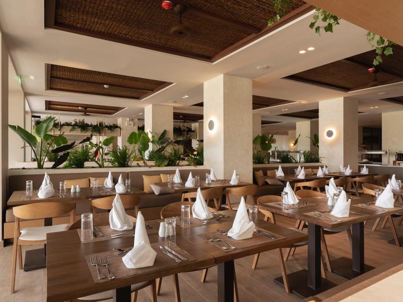 Großer, heller Speisesaal mit vielen Holztischen, weißen Servietten und grünen Pflanzen.